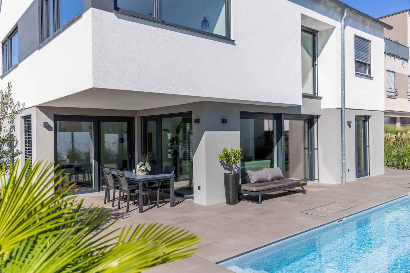 Modern white house with large windows, patio, dining table, lounge chair, and a narrow swimming pool surrounded by plants.