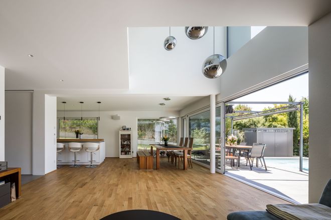 Modern open-plan living area with wooden floors, a kitchen, dining table, and floor-to-ceiling windows leading to a patio and pool.