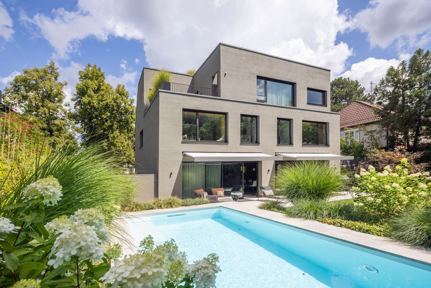 Modern three-story gray house with large windows, surrounded by lush greenery and featuring a rectangular swimming pool in the backyard.