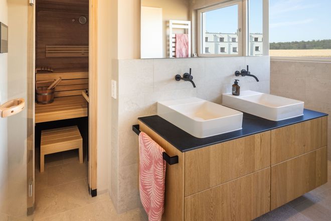 Modern bathroom with twin sinks on a wooden vanity, black faucets, towel, large mirror, and a sauna with wooden seating and a bucket inside.