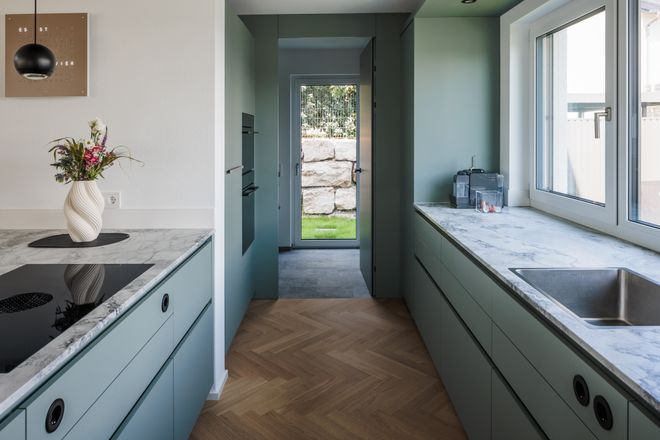 Modern kitchen with mint green cabinets, marble countertops, and a vase of flowers. Herringbone wood floor leads to a glass door opening to a garden.