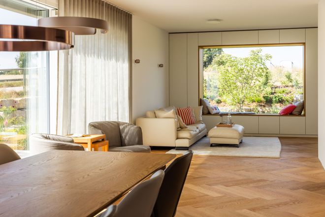 Modern living room with large window, white sofa, wooden dining table, and chevron-patterned floor. View of garden outside.