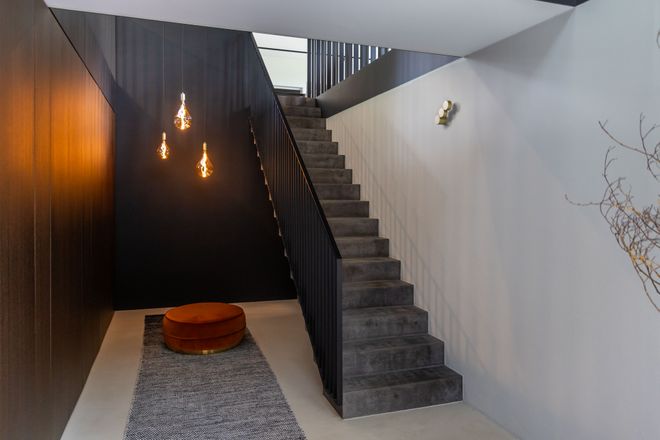 Minimalist interior with a staircase, dark wall, hanging lights, a gray rug, and a small round orange seat, creating a modern aesthetic.