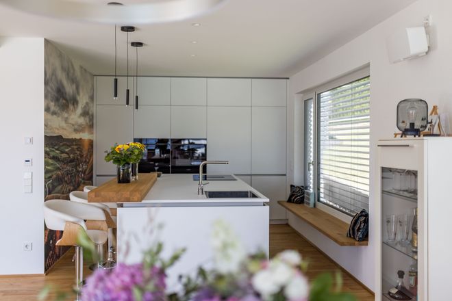 Modern kitchen with white cabinets, island with wooden countertop, bar stools, and large window. Vase with sunflowers on the island.
