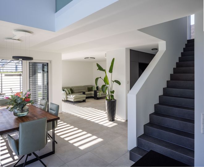 Modern interior with a staircase, dining table, and a gray L-shaped sofa. Large windows with blinds cast striped shadows on the floor.