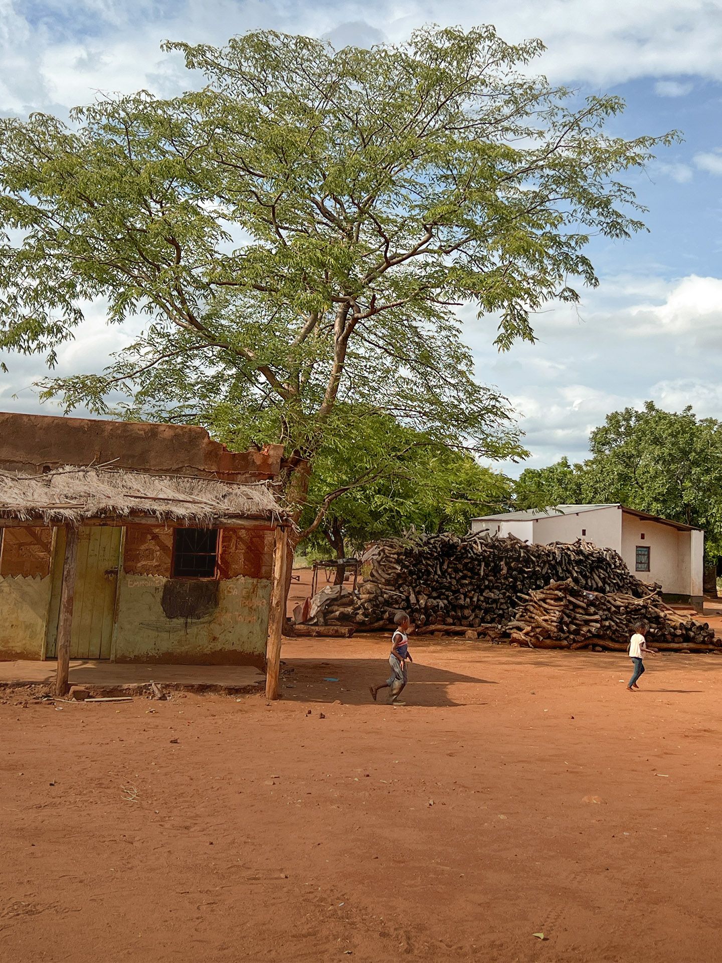 Afrikanische Huette auf rotem Sand davor spielende Kinder im Hintergrund blauer Himmel und Baeume
