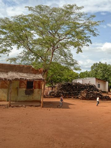 Afrikanische Huette auf rotem Sand davor spielende Kinder im Hintergrund blauer Himmel und Baeume