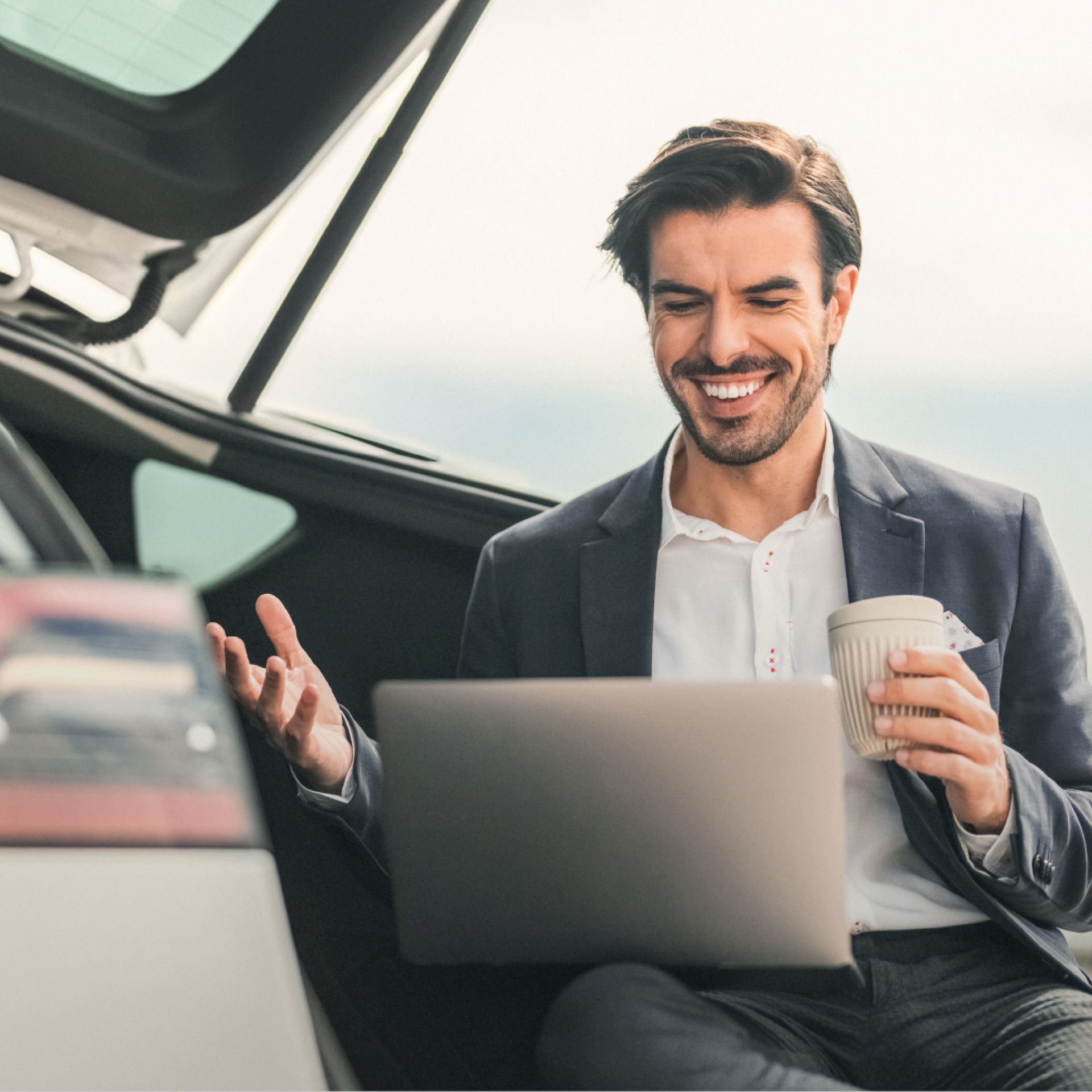 Mann lädt sein E-Auto auf und arbeitet währenddessen am Laptop mit Kaffee in der Hand