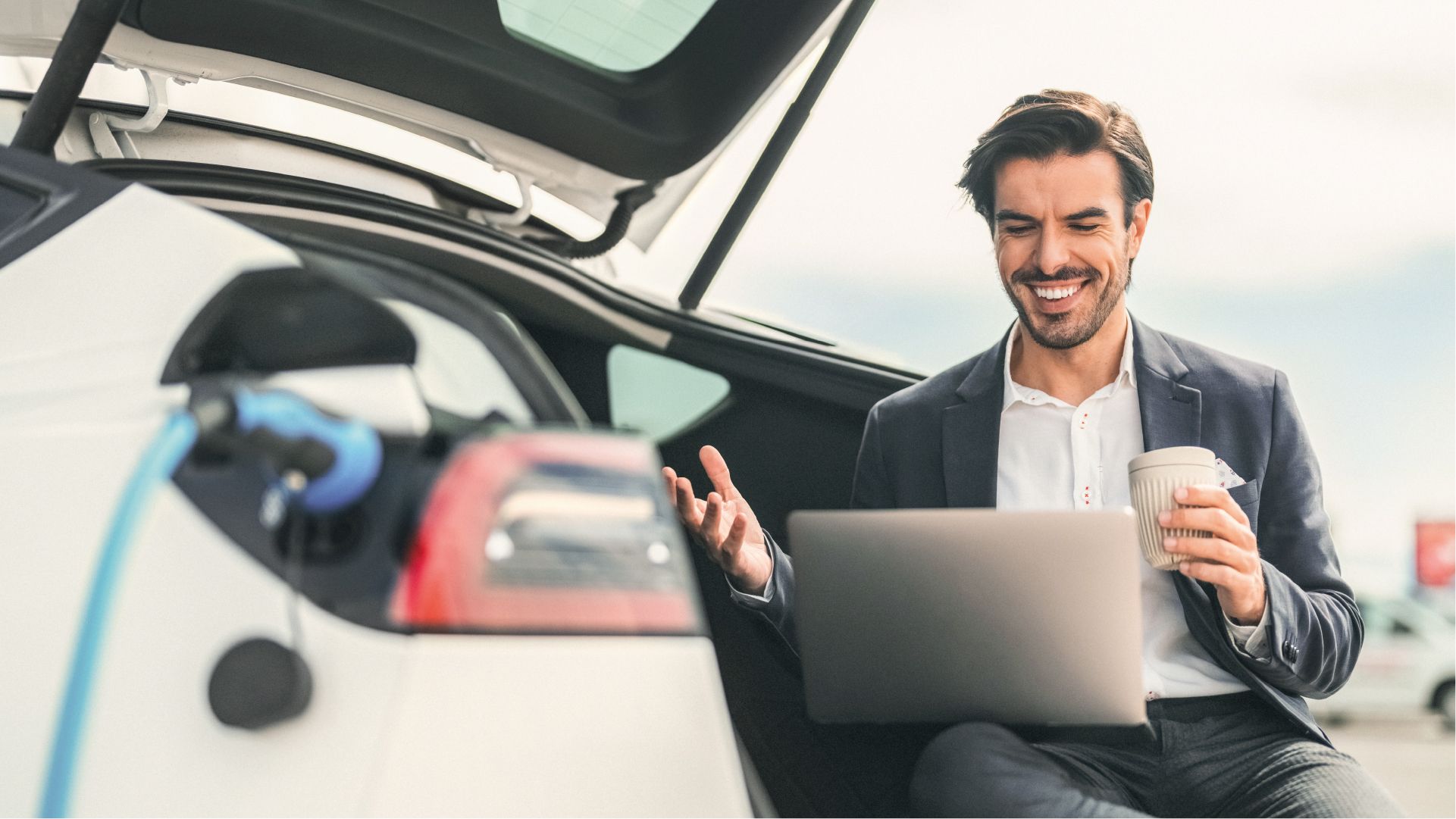 Mann lädt sein E-Auto auf und arbeitet währenddessen am Laptop mit Kaffee in der Hand