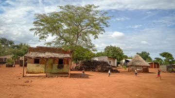 Afrikanische Huette auf rotem Sand davor spielende Kinder im Hintergrund blauer Himmel und Baeume