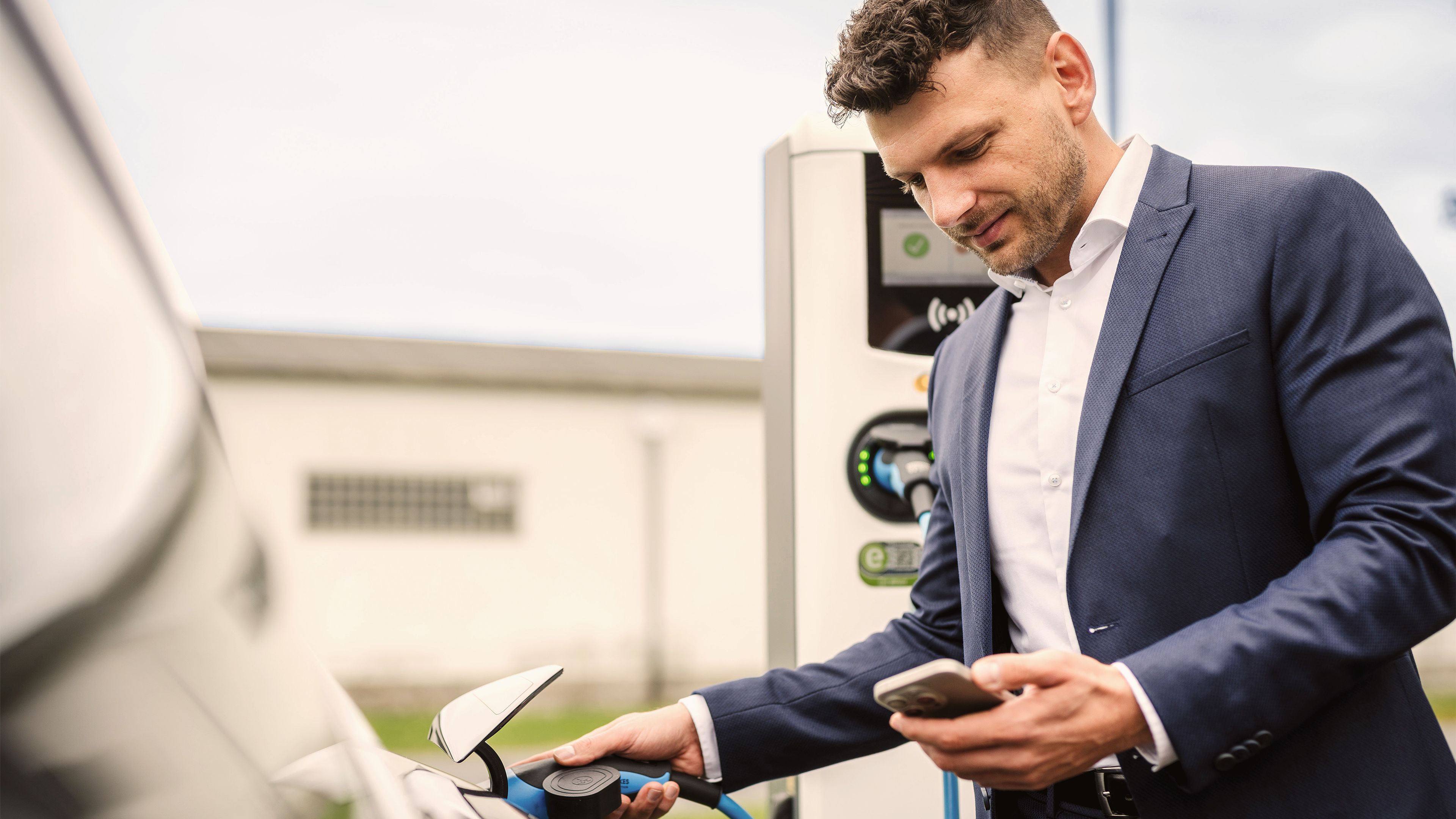 Mann im Anzug laedt E-Auto und schaut dabei aufs Handy