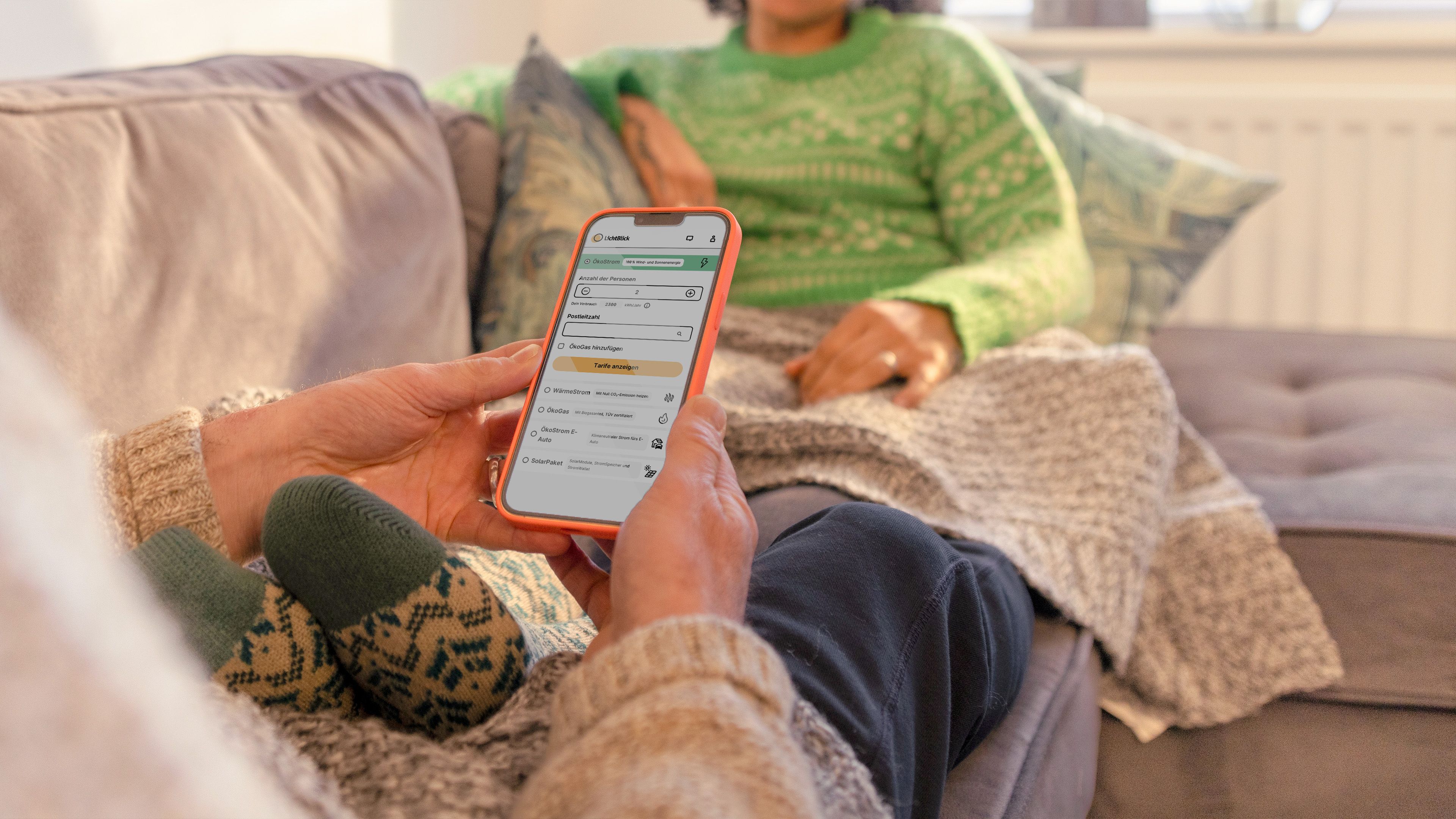 Zwei Personen sitzen gemütlich auf dem Sofa, eine Person hat ein Handy mit dem LichtBlick Stromrechner in der Hand