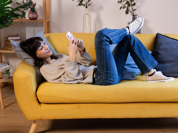 Frau am Handy liegt auf einem gelben Sofa
