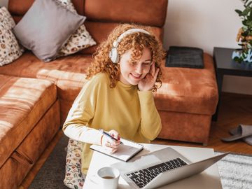 Frau mit roten Locken sitzt mit Kopfhoerern am Laptop und schreibt etwas