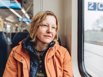 Frau mit orangener Jacke sitzt in der Bahn und guckt aus dem Fenster