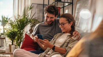 Paar sitzt im Wohnzimmer auf der Couch und guckt aufs Handy