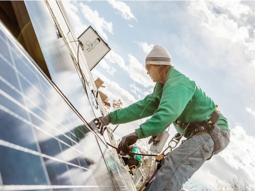 Ein Installateur mit grünem Sweatshirt, Sonnenbrille und weißer Mütze montiert Solarmodule auf einem steilen Hausdach.