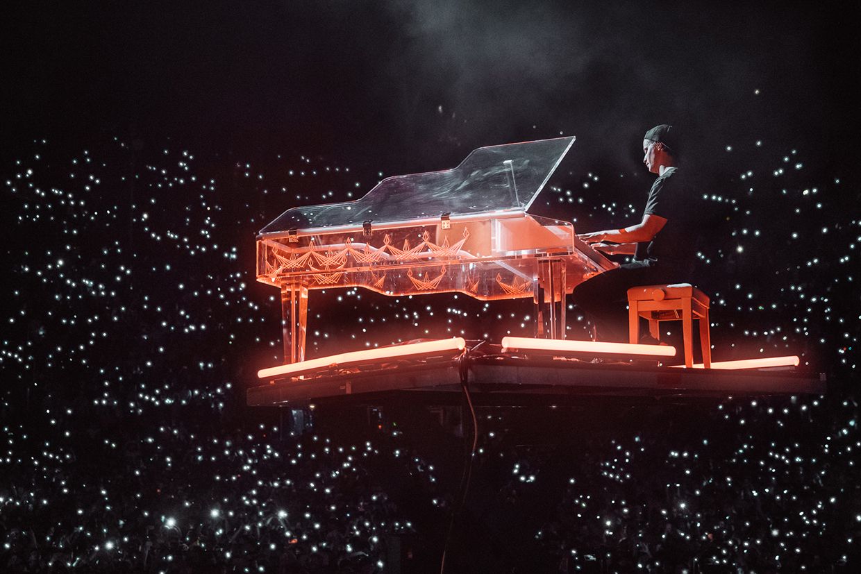 Musician playing a transparent grand piano on a lit platform, surrounded by a sea of lights from the audience in a darkened venue.