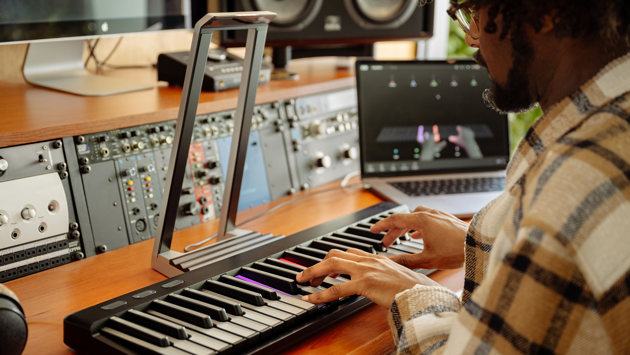 Person playing a digital keyboard in a studio, with audio equipment and a laptop displaying a virtual instrument in the background.