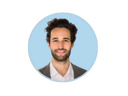 Smiling man with curly hair and beard, wearing a white shirt and gray blazer, against a light blue background.