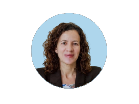 Woman with curly hair in a brown blazer, smiling against a light blue background.
