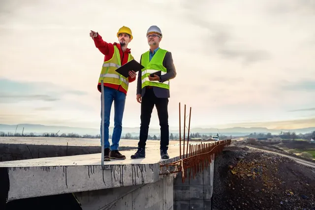 Two construction workers in safety gear on a bridge, discussing plans, with one pointing towards the horizon.