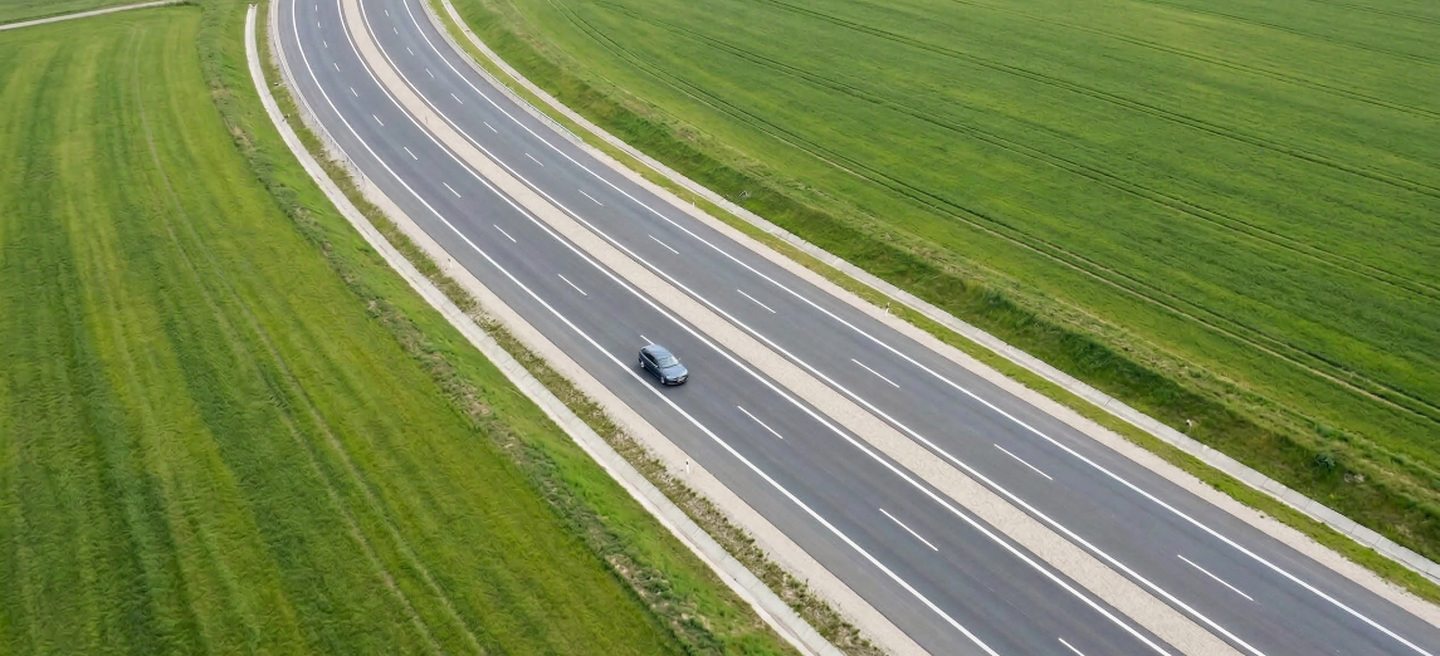 A car travels on a winding highway through expansive green fields, captured from an aerial perspective.