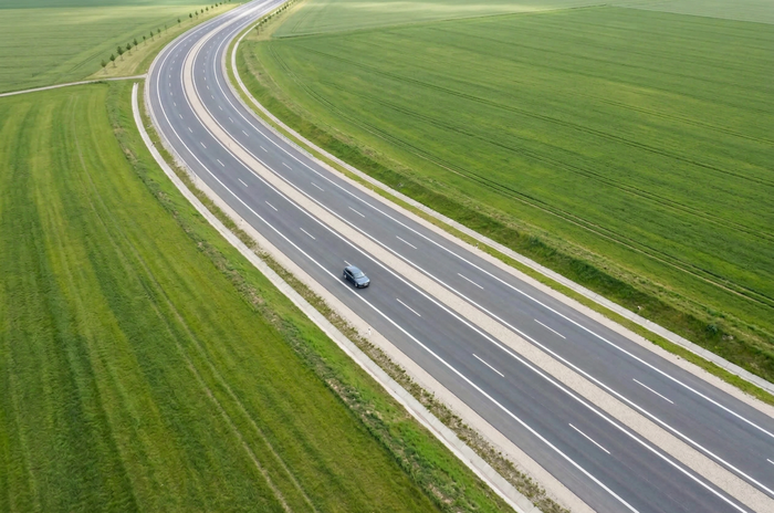 A car travels on a winding highway through expansive green fields, captured from an aerial perspective.