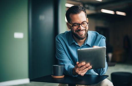 Smiling man with glasses uses a tablet at a table with a coffee mug, indoors.