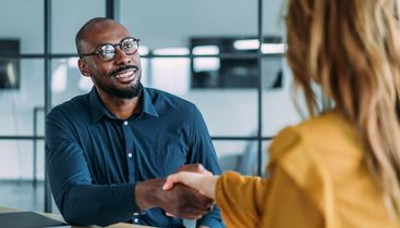 Man in glasses smiling and shaking hands with a woman in an office setting, demonstrating a professional interaction.