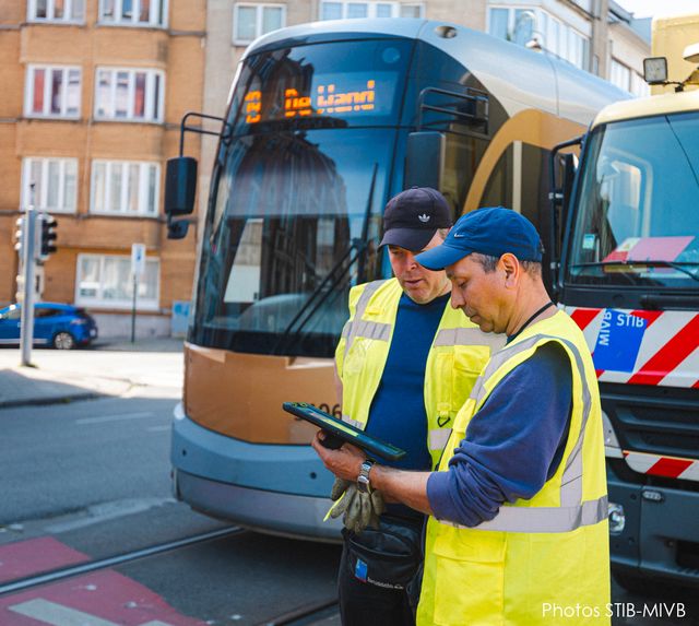 Two STIB-MIVB maintenance workers wearing high-visibility safety vests review information on a tablet beside a modern tram and a service vehicle in an urban street.