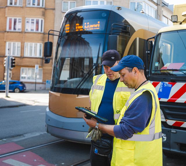 Two STIB-MIVB maintenance workers wearing high-visibility safety vests review information on a tablet beside a modern tram and a service vehicle in an urban street. The scene highlights field operations, real-time asset management, and public transport infrastructure maintenance in Brussels.