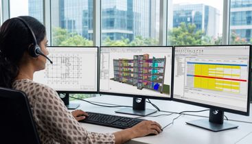 A person with a headset works at a desk using three monitors displaying architectural designs and data spreadsheets.