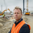 Homme en gilet orange regardant directement la caméra sur un site industriel avec des grues