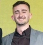 Smiling man in a gray blazer and dark shirt against a yellow background.