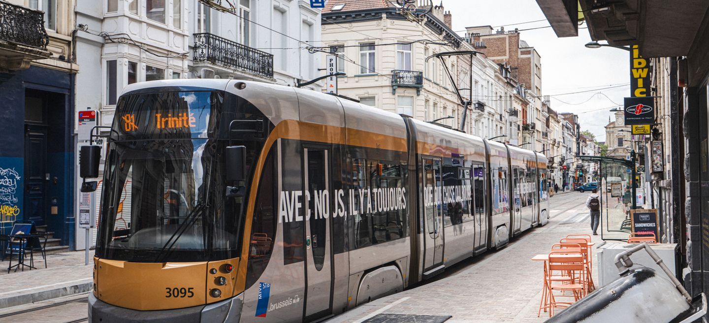 A modern STIB-MIVB tram travels through a narrow Brussels city street lined with historic buildings, shops, and cafés.