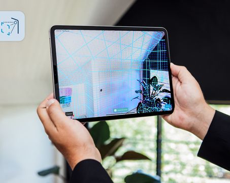 Person holding a tablet displaying a 3D scanned room with a grid overlay, showcasing augmented reality technology.