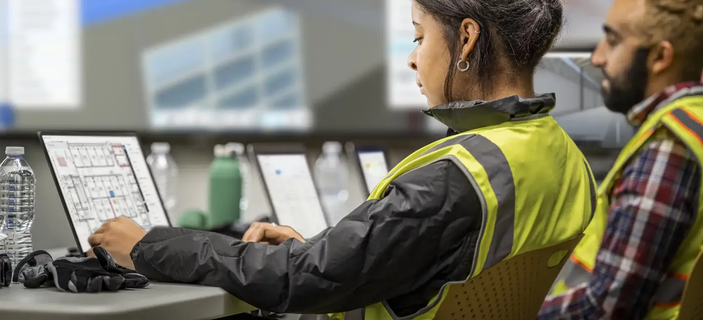 A young woman and a man collaborate at a workstation using Autodesk software during an onboarding session, reviewing project tools and workflows on a large screen in a modern office environment