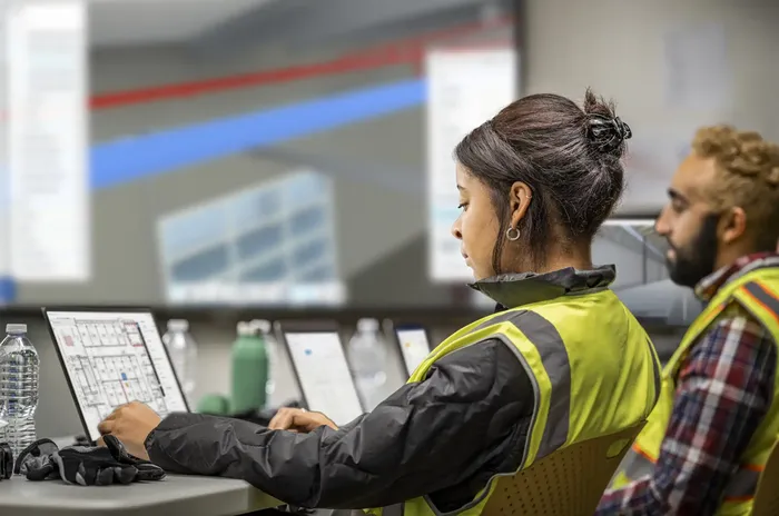 A young woman and a man collaborate at a workstation using Autodesk software during an onboarding session, reviewing project tools and workflows on a large screen in a modern office environment