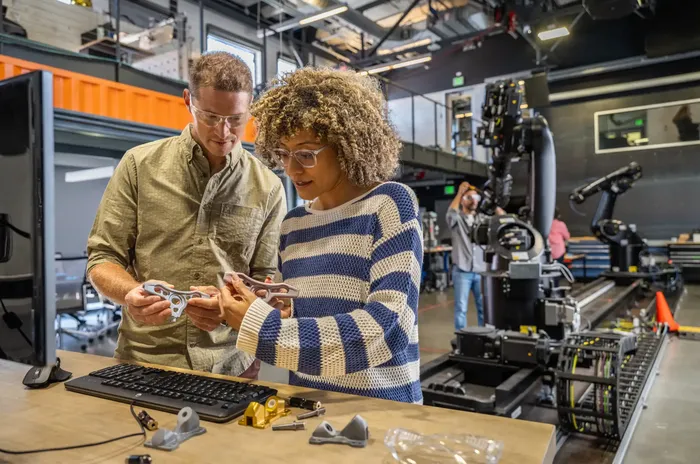 Two engineers collaborate on a manufacturing floor, using engineering software to review product design and production details.