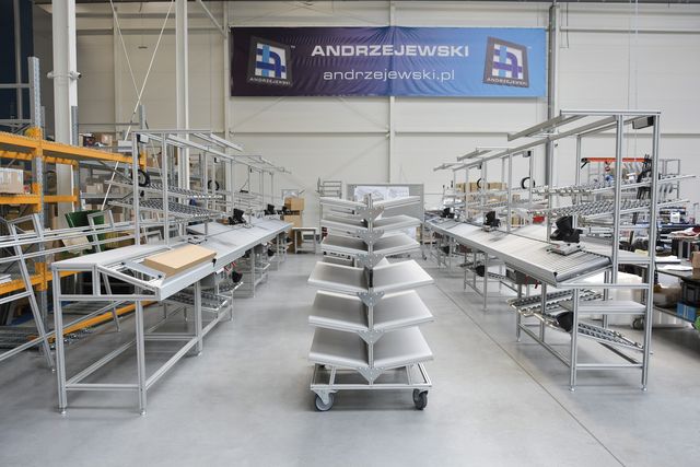 Industrial workshop with metal racks and assembly lines, under a large "Andrzejewski" banner in a spacious, well-lit factory setting.