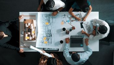 Aerial view of a team collaborating over architectural blueprints, a model, and a laptop on a table, with notes and coffee cups scattered around.