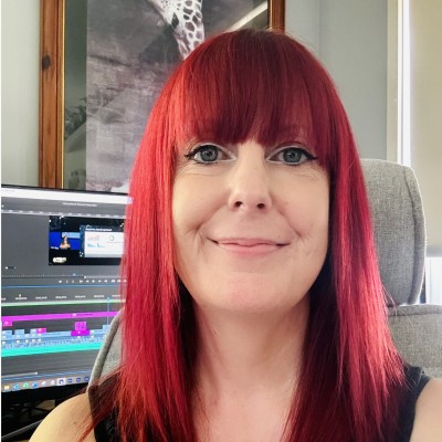 Person with red hair smiling, sitting in front of a computer with video editing software on the screen, in a cozy room.