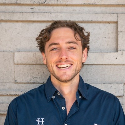 Smiling man with wavy hair and wearing a navy blue button-up shirt stands against a textured stone wall.