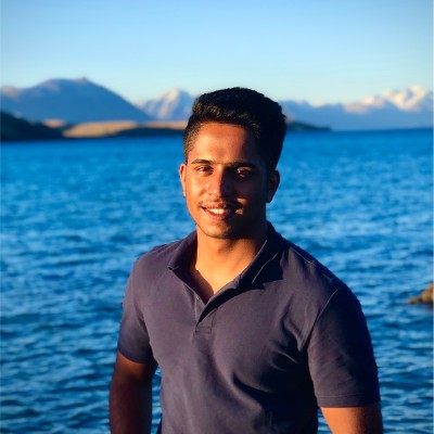 A person smiling by a lake with mountains in the background under a clear blue sky.