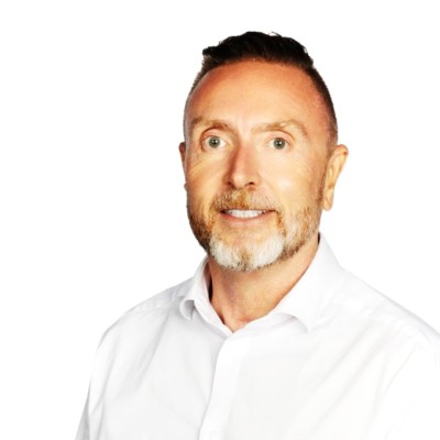 A man with short hair and a beard, wearing a white shirt, smiles against a plain white background.