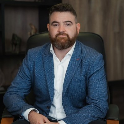 Bearded man in a blue suit and white shirt sits in a black chair, with a blurred office background.