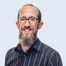 Smiling man with glasses and a beard, wearing a striped shirt, against a light blue background.