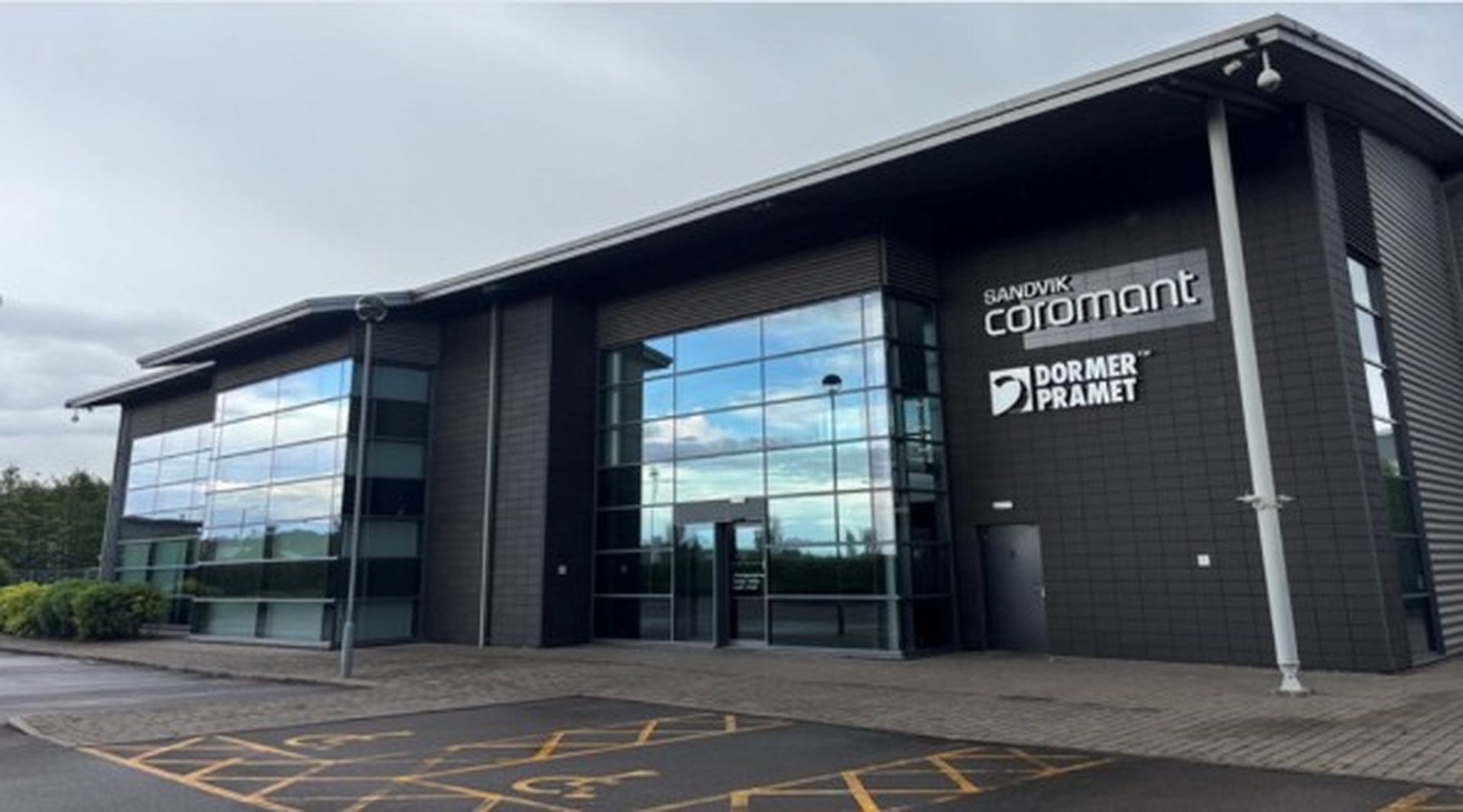 Modern building with reflective glass facade displaying "Sandvik Coromant" and "Dormer Pramet" logos, under a cloudy sky.