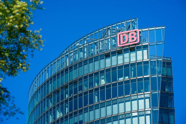 Glass office building with a red "DB" logo against a clear blue sky, partially framed by green foliage.